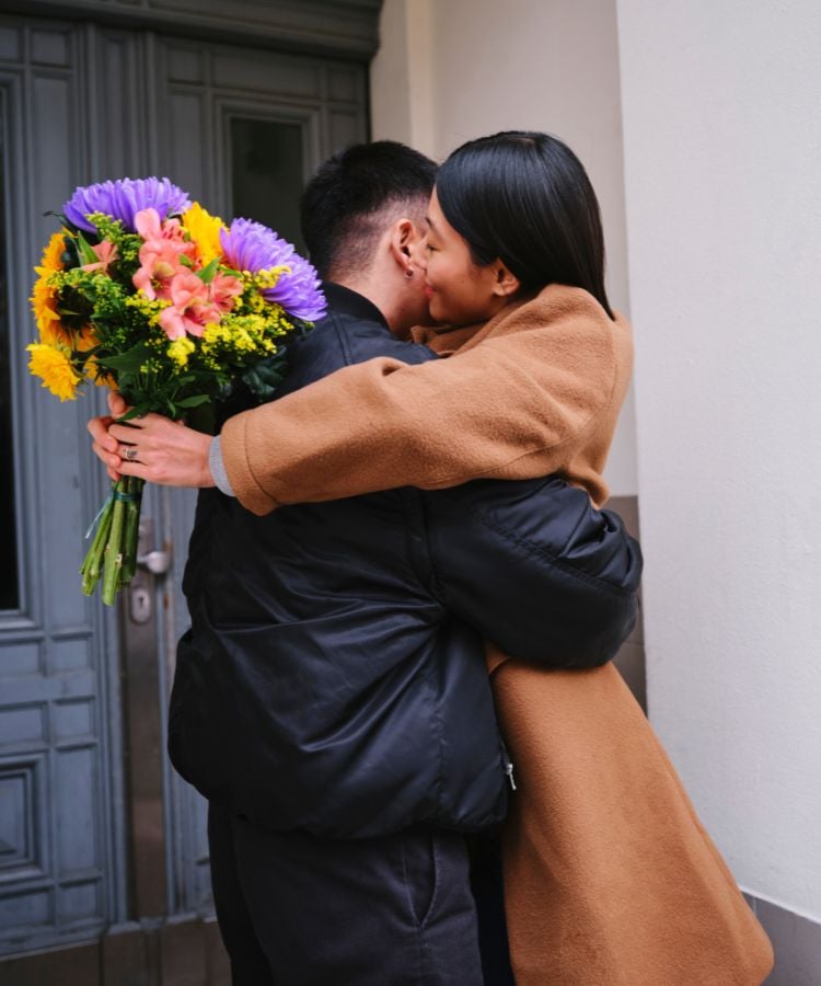 Um casal está se abraçando em frente a uma porta. A mulher, que está de costas para a câmera, segura um buquê de flores coloridas. Ela está usando um casaco marrom, enquanto o homem, que está de frente para a câmera, veste um casaco preto. Eles parecem estar felizes e emocionados.