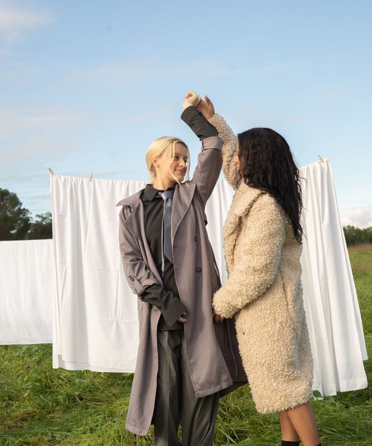 Duas mulheres dançam em um campo com lençóis brancos pendurados ao fundo. Uma delas tem cabelo loiro e veste um casaco cinza, a outra tem cabelo escuro e veste um casaco bege.