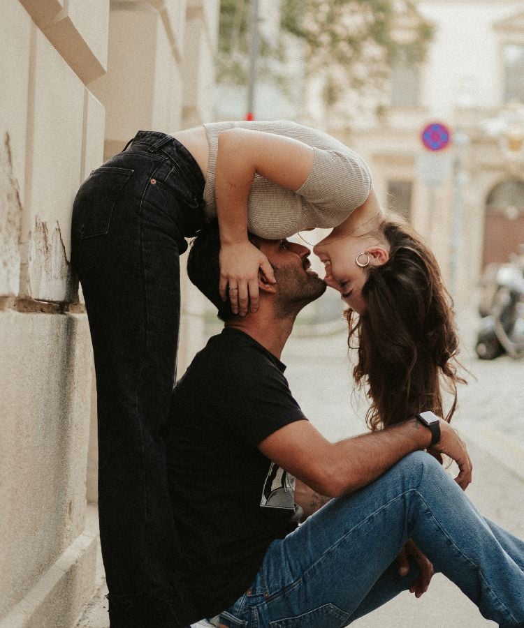 Um casal está em um momento romântico na rua. O homem está sentado no chão, encostado em uma parede, com as pernas dobradas. A mulher está de pé, inclinada para frente, com as mãos apoiadas nos ombros do homem, e eles estão prestes a se beijar. A mulher está usando uma blusa curta e calça jeans, enquanto o homem veste uma camiseta preta e calça jeans.