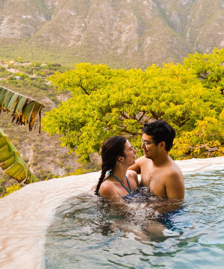 Um casal está em uma piscina infinita, cercada por montanhas e vegetação exuberante. Eles se olham carinhosamente, com sorrisos nos rostos. A mulher usa um biquíni azul e o homem está sem camisa.