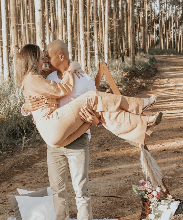 Um homem carrega sua namorada nos braços em uma floresta de árvores altas. Eles se olham com amor e sorrisos. A mulher veste um conjunto bege e o homem uma camisa branca e calça cinza.