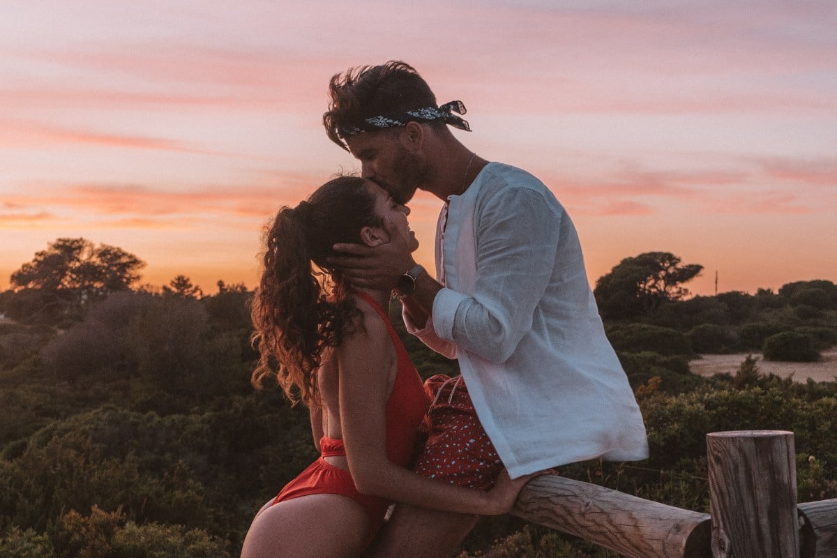 A imagem mostra um casal em um momento romântico ao ar livre durante o pôr do sol. A mulher está vestindo um maiô vermelho e tem cabelos cacheados presos em um rabo de cavalo. O homem está usando uma camisa branca de mangas compridas e uma bandana preta na cabeça. Ele segura o rosto da mulher com ambas as mãos e a beija na testa. Eles