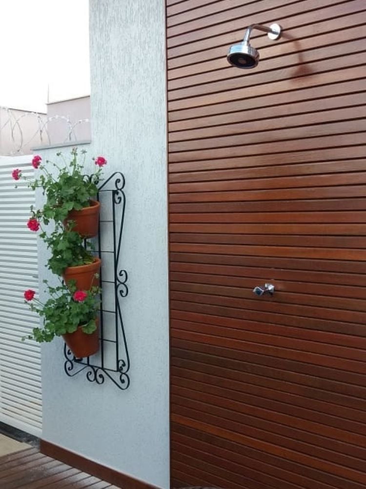 Foto de suporte preto com vasos de gerânios pendentes vermelhos, ao lado de ducha no terraço