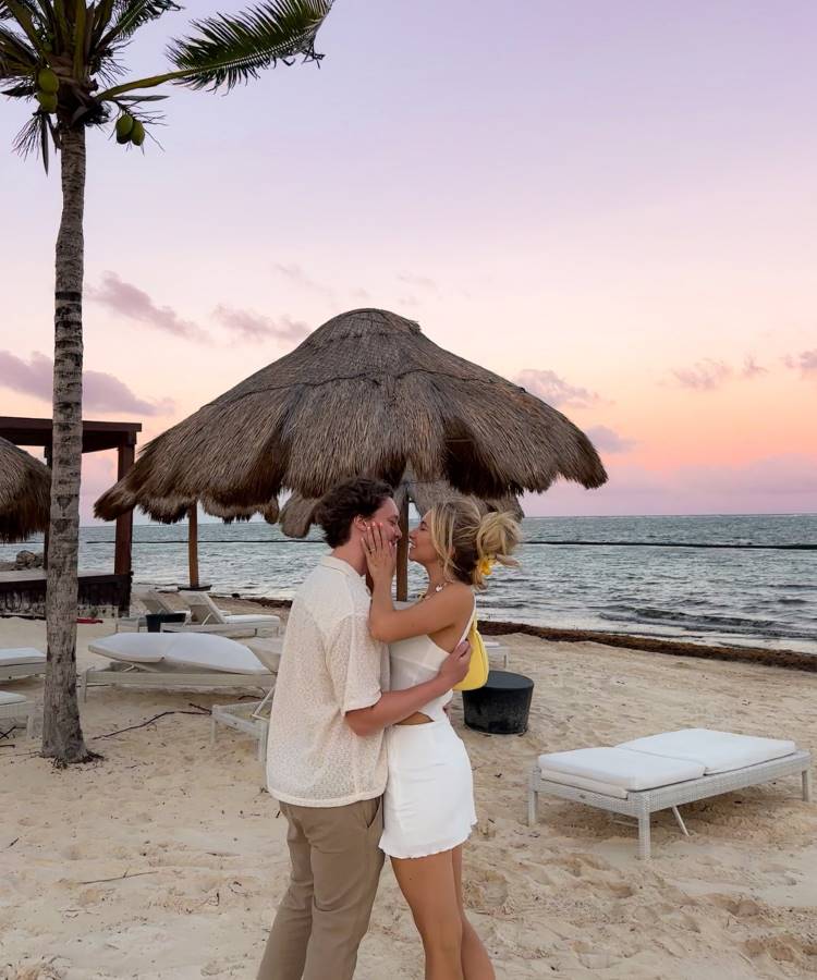 Um casal se abraça na praia ao pôr do sol. A mulher, de saia branca e top, toca o rosto do homem, que veste uma camisa clara. Ao fundo, há guarda-sóis de palha e o mar. Fotos tumblr na praia frequentemente capturam momentos românticos como este.