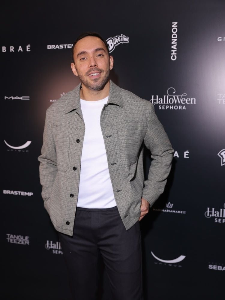 A imagem mostra um homem sorrindo em frente a um painel de marcas. Ele tem cabelo curto e barba. Veste uma jaqueta cinza sobre uma camiseta branca e calças pretas. O fundo é preto com logotipos de várias marcas em branco, incluindo "Halloween da Sephora 2024".