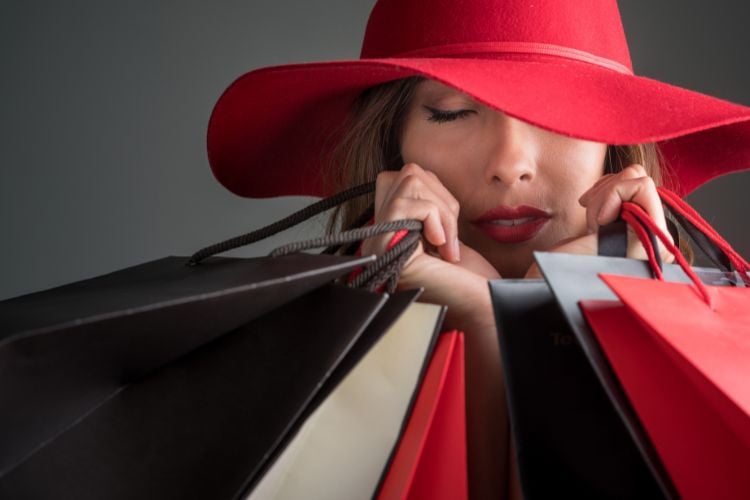 A imagem mostra uma mulher usando um chapéu vermelho elegante, com os olhos fechados e um leve sorriso. Ela está cercada por várias sacolas de compras em tons de vermelho e preto. A cena transmite uma sensação de satisfação e estilo. Essa imagem exemplifica como a inteligência artificial pode ajudar a criar o look perfeito para um dia de compras.