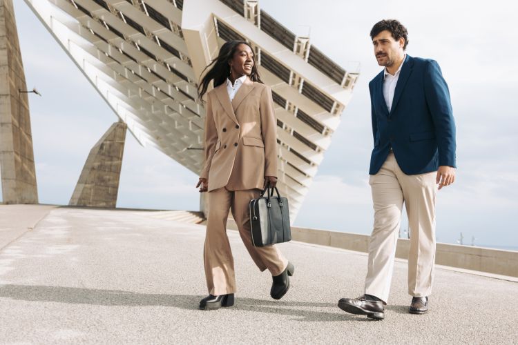 A imagem mostra duas pessoas caminhando lado a lado em um ambiente urbano. À esquerda, uma mulher veste um terno bege e segura uma bolsa preta. Ela está sorrindo e parece estar em movimento. À direita, um homem veste um blazer azul, camisa branca e calças claras. Ao fundo, há uma estrutura arquitetônica moderna, que parece ser parte de um edifício ou ponte. O cenário é ao ar livre, sob um céu claro.