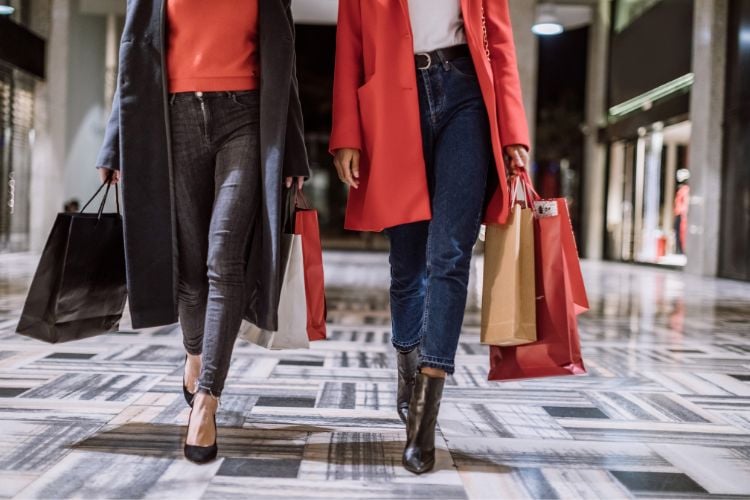 A imagem mostra duas pessoas caminhando em um ambiente de compras. Ambas estão segurando várias sacolas de compras. A pessoa à esquerda veste um casaco escuro e calça jeans, enquanto a pessoa à direita usa um casaco vermelho e botas pretas. O chão tem um padrão geométrico elegante. Essa cena poderia ser um exemplo de como a inteligência artificial ajuda a criar o look perfeito para um dia de compras.