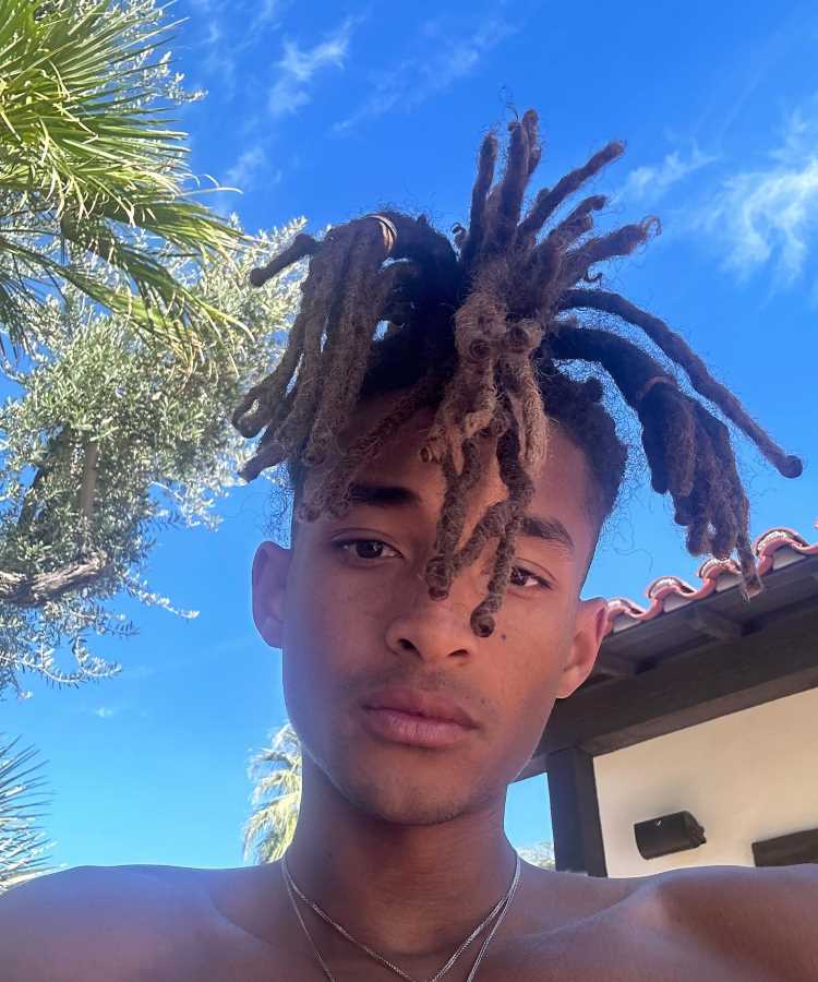 A imagem mostra um jovem, Jaden Smith, com dreadlocks longos e volumosos. Ele está ao ar livre, sob um céu azul claro. Ao fundo, há palmeiras e parte de um telhado de telhas. O jovem está sem camisa e usa um colar fino. A luz do sol ilumina seu rosto.