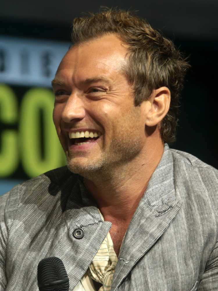 A imagem mostra Jude Law sorrindo, segurando um microfone. Ele está vestindo um casaco cinza com listras finas e uma camisa clara por baixo. O fundo parece ser de um evento, possivelmente uma convenção, com letras desfocadas. O homem tem cabelo curto e barba por fazer.