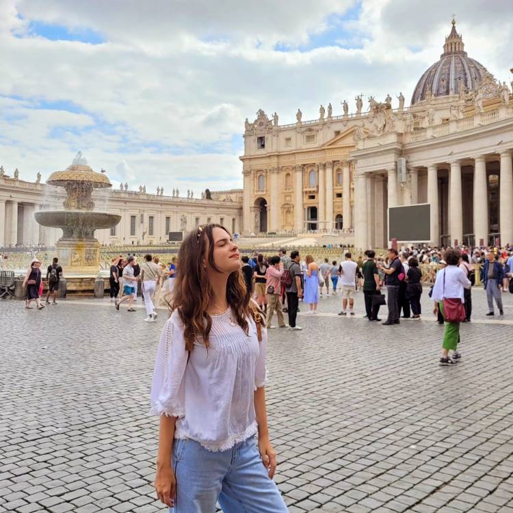 A imagem mostra uma mulher em pé na Praça de São Pedro, no Vaticano. Ela está vestindo uma blusa branca e calças jeans, com os olhos fechados, parecendo apreciar o momento. Ao fundo, há uma fonte e a fachada da Basílica de São Pedro, com várias pessoas ao redor, sugerindo um ambiente turístico. O céu está parcialmente nublado.