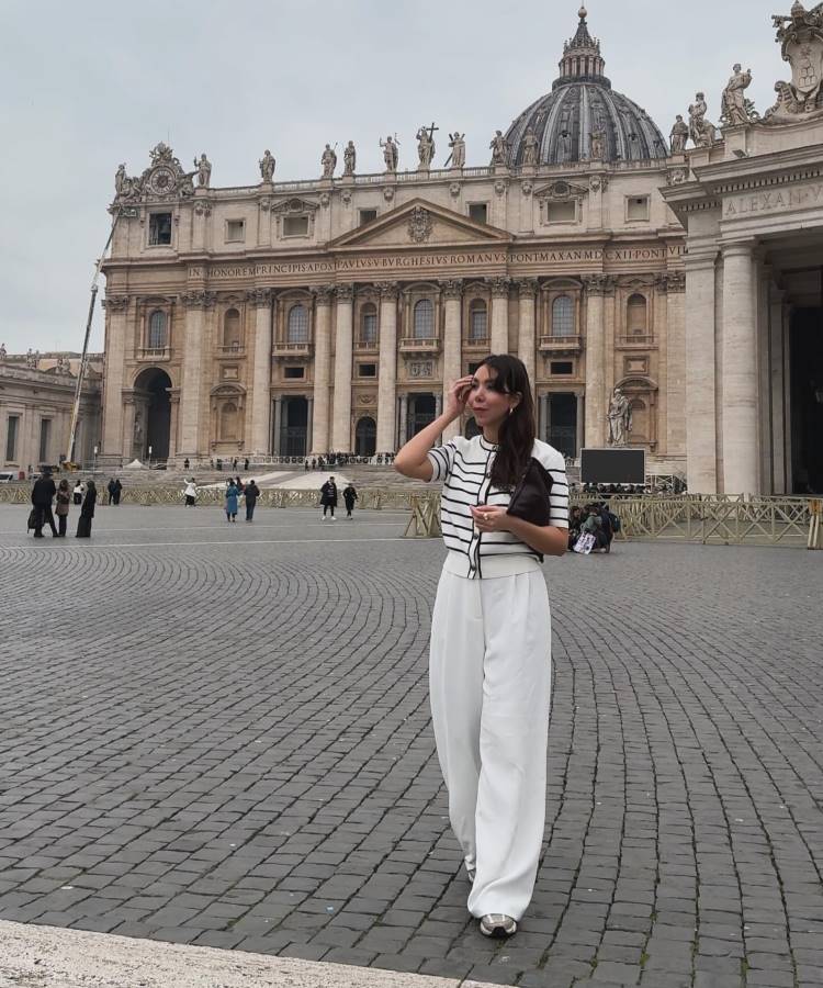 A imagem mostra uma mulher em pé na Praça de São Pedro, no Vaticano. Ela está vestindo uma blusa listrada em preto e branco e calças brancas largas. A mulher segura uma bolsa pequena e parece estar ajustando o cabelo. Ao fundo, está a fachada da Basílica de São Pedro, com sua arquitetura imponente e detalhada. O céu está nublado, e poucas pessoas estão espalhadas pela praça.