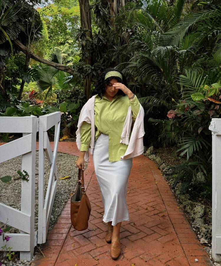 Uma mulher caminha por um caminho de tijolos em um jardim tropical. Ela usa uma blusa verde clara de mangas longas, saia branca longa e sapatos marrons. Um suéter rosa está jogado sobre os ombros, e ela segura uma bolsa marrom grande. Ela usa uma faixa verde no cabelo.