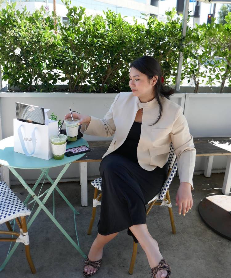 Uma mulher está sentada em um café ao ar livre, cercada por plantas. Ela veste um vestido preto longo e um blazer bege. Com sandálias marrons, segura uma bebida verde em um copo de plástico. Há uma mesa verde clara com mais bebidas e uma bolsa ao lado.