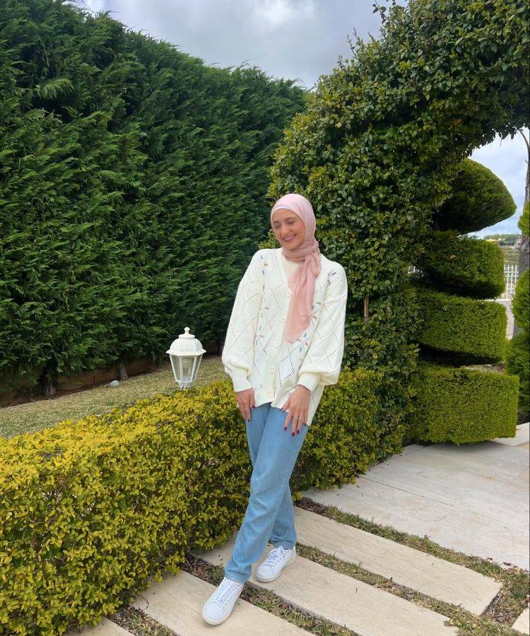 Em um jardim bem cuidado, uma mulher veste um suéter branco de tricô, jeans azul claro e tênis brancos. Ela usa um hijab rosa claro. Este estilo é um exemplo de moda modesta.