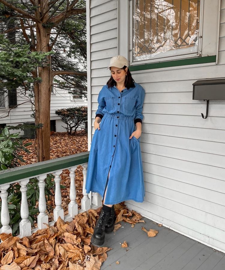 Numa varanda com folhas secas, uma mulher veste um vestido jeans longo, azul, de mangas compridas, e botas pretas. Ela usa um boné bege e está encostada na parede. Este visual é um exemplo de moda modesta.