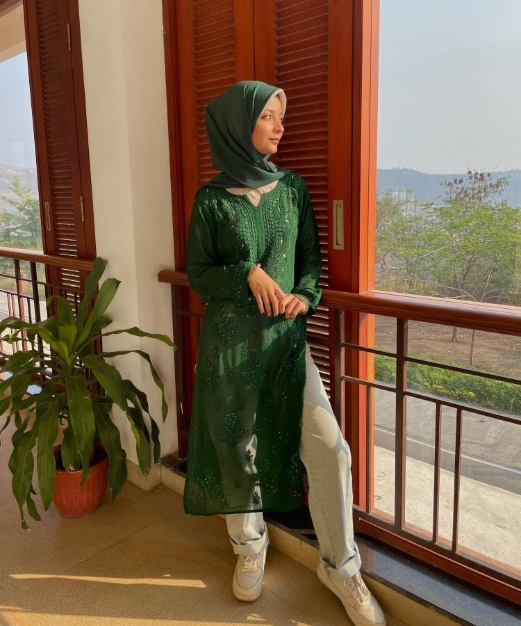 Em uma varanda ensolarada, uma mulher usa um vestido longo verde com bordados, jeans claro e tênis brancos. Ela complementa o visual com um hijab verde. Este estilo é um exemplo de moda modesta.