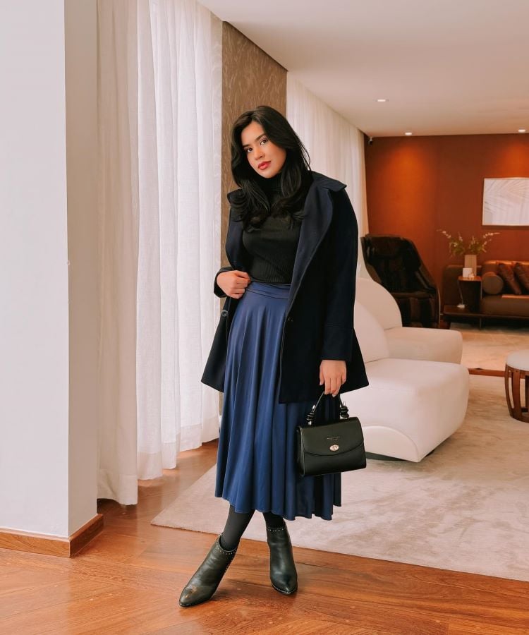 Em uma sala elegante, uma mulher usa um suéter preto, saia azul plissada até os tornozelos, casaco preto e botas pretas. Ela segura uma bolsa preta. Este look representa a moda modesta.