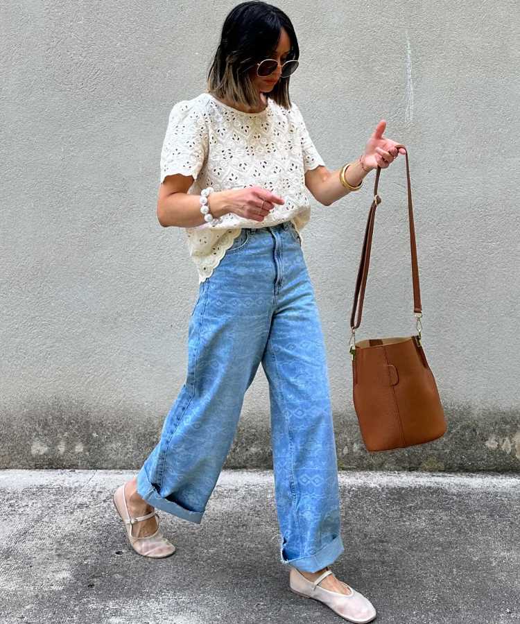 A imagem mostra uma mulher vestindo uma blusa de renda branca com detalhes vazados, combinada com uma calça jeans de corte largo. Ela usa sapatilhas bege e segura uma bolsa marrom de alça longa. Óculos de sol completam o visual. Este look é casual e elegante, adequado para ir à missa em um ambiente mais descontraído.