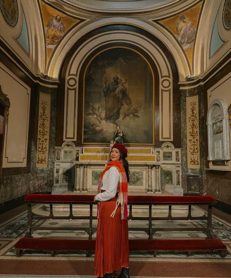 A imagem mostra uma mulher em uma igreja, vestindo um conjunto elegante e apropriado para ir à missa. Ela usa uma saia longa plissada em tom terracota, combinada com um suéter branco. Um cachecol listrado em tons de vermelho e laranja complementa o visual, junto com uma boina vermelha. Ao fundo, há um altar com uma pintura religiosa e estátuas, realçando o ambiente sagrado.