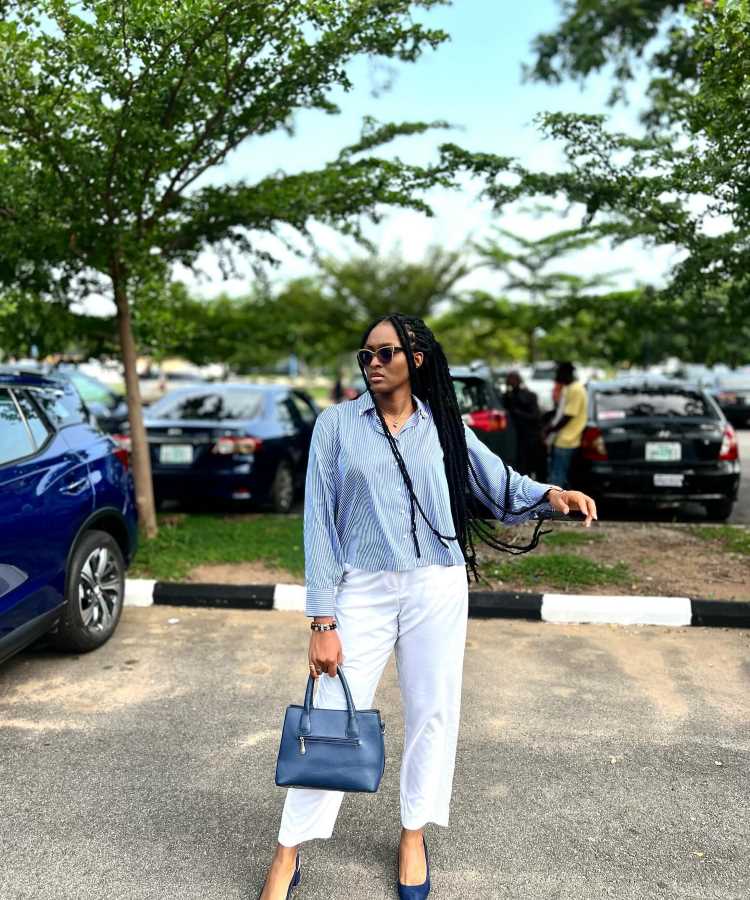 A imagem mostra uma mulher em um estacionamento ao ar livre, cercada por árvores. Ela está vestindo uma camisa listrada azul e branca, combinada com calças brancas. Ela segura uma bolsa azul e usa sapatos de salto na mesma cor. Seus cabelos estão trançados e ela usa óculos escuros. Ao fundo, há vários carros estacionados.