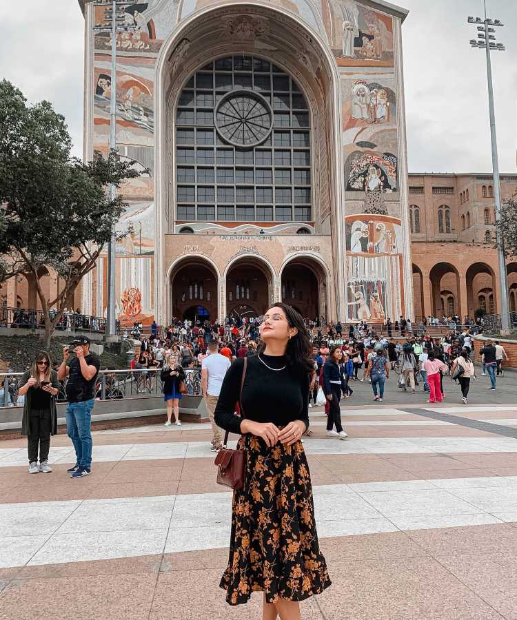 A imagem mostra uma mulher em frente à Basílica de Nossa Senhora Aparecida. Ela usa um suéter preto e uma saia midi com estampa floral em tons de marrom e dourado. Completa o look com uma bolsa marrom e óculos de sol. Este é um look elegante e apropriado para ir à missa em um lugar tão significativo. Ao fundo, há muitas pessoas e a grandiosa fachada da basílica.