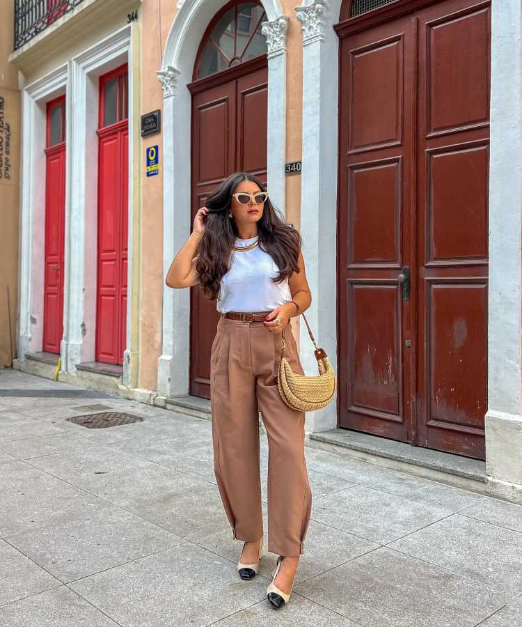 A imagem mostra uma mulher em uma rua com portas vermelhas ao fundo. Ela está vestindo uma blusa branca sem mangas e calças de cintura alta em tom bege. Usa óculos de sol brancos e segura uma bolsa de palha. Nos pés, calça sapatos de bico preto e salto baixo. O cabelo está solto e ela parece estar caminhando ou posando para a foto.