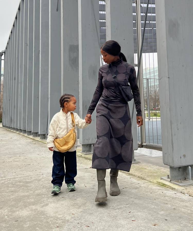 A imagem mostra uma mulher e uma criança caminhando de mãos dadas em um ambiente urbano. A mulher está vestida com um vestido longo de mangas compridas com estampa circular e botas. Ela carrega uma pequena bolsa preta transversal. A criança usa uma jaqueta clara, calças escuras e tênis verdes, com uma bolsa amarela no ombro. Eles estão em frente a uma estrutura metálica cinza, e o clima parece estar nublado. A imagem mostra uma mulher em pé em um grande salão com piso de mármore decorado. Ela está vestida com roupas claras e um cachecol, e carrega uma bolsa transversal. Ao fundo, há um ambiente imponente com colunas e detalhes arquitetônicos elaborados. Várias pessoas estão ao fundo, sugerindo que o local está aberto ao público, possivelmente um local turístico ou histórico.