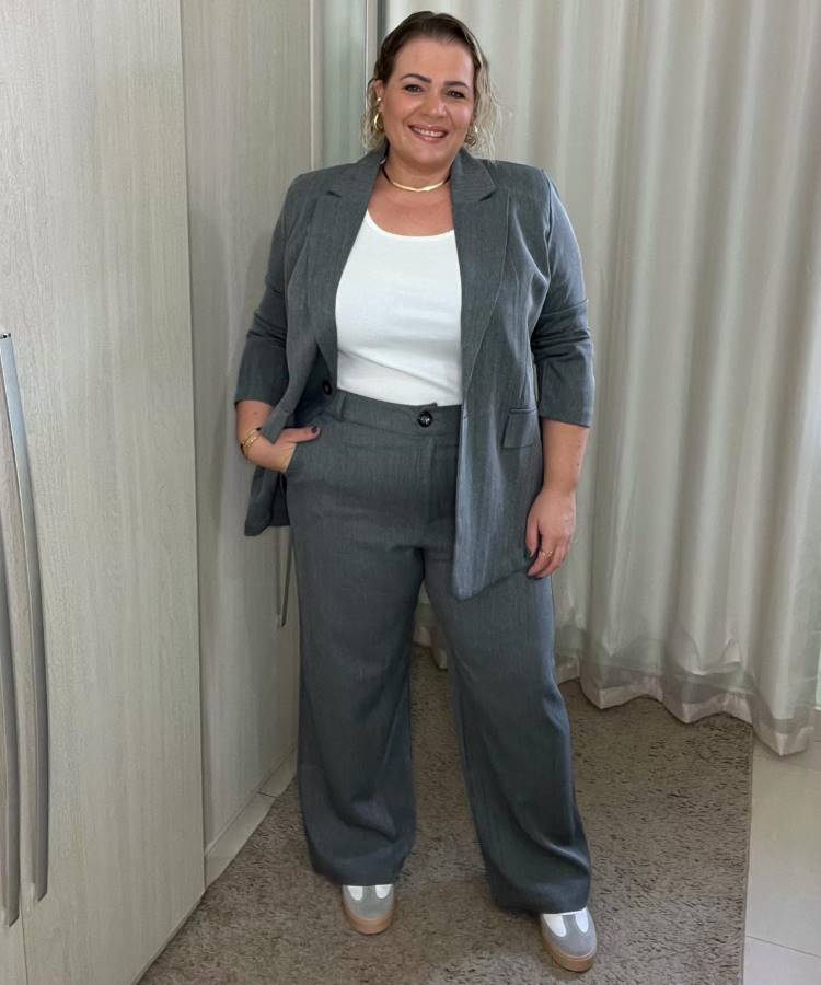 A imagem mostra uma mulher sorridente vestindo um conjunto de terno cinza com calças largas e um blazer. Ela combina o terno com uma blusa branca e sapatos de plataforma brancos e dourados. Este é um dos looks para visitar igrejas, pois é elegante e apropriado para ambientes mais formais. Ela está em um espaço interno com cortinas claras ao fundo.