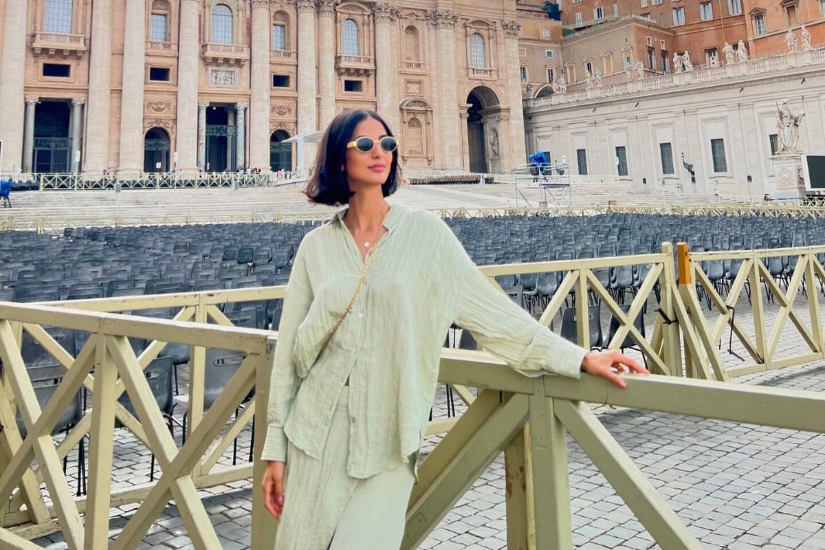 A imagem mostra uma mulher em frente a uma igreja monumental, provavelmente a Basílica de São Pedro, no Vaticano. Ela está vestindo um conjunto de blusa e saia longa de tecido leve em tom claro, ideal para o clima e para a ocasião. Ela usa óculos de sol e carrega uma bolsa transversal. Este é um dos looks para visitar igrejas, pois é elegante e respeitoso, adequado para locais religiosos. O ambiente ao redor é composto de cadeiras e uma estrutura arquitetônica grandiosa.