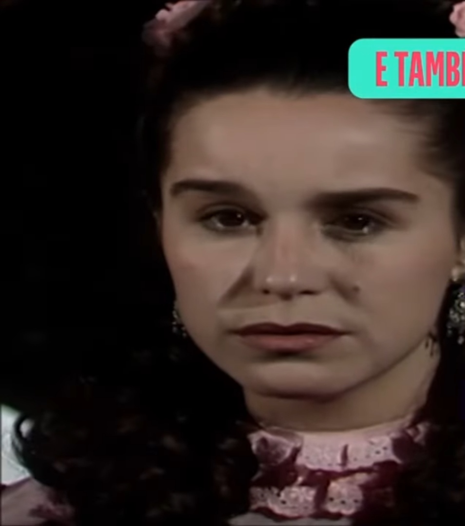 Lucélia Santos em uma cena da novela "Escrava Isaura" dos anos 80, onde interpretou a personagem principal. Ela está com expressão séria, usando um vestido de época com detalhes rendados. A imagem destaca sua atuação marcante e o impacto cultural da novela na televisão brasileira.