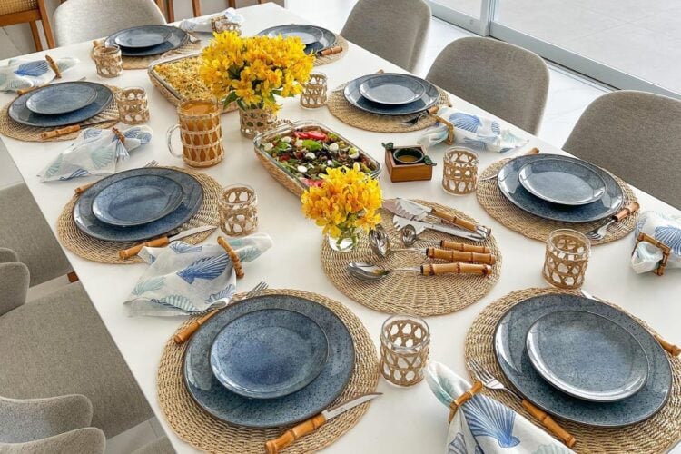 A imagem mostra uma mesa posta para refeição, decorada de forma elegante. Cada lugar tem sousplats de palha, pratos de cerâmica azul e talheres com cabos de bambu. Guardanapos brancos com estampas azuis estão presos por anéis de madeira. Copos transparentes têm detalhes de palha. No centro, há arranjos de flores amarelas vibrantes, uma travessa com salada colorida e um prato de forno. A mesa combina elementos naturais e rústicos, criando um ambiente acolhedor e sofisticado.