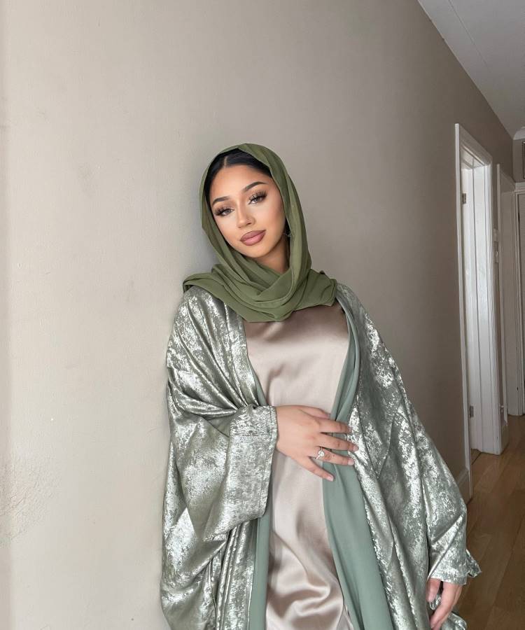 Uma mulher está encostada em uma parede interna. Ela veste um vestido longo de seda bege com um kimono verde brilhante e um hijab verde combinando. Este estilo elegante é um exemplo de moda modesta.