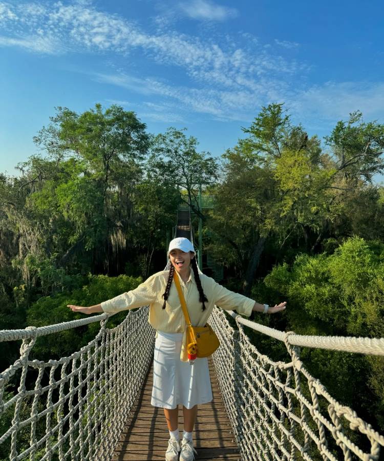 Uma mulher está em uma ponte suspensa cercada por árvores. Ela usa uma blusa amarela de mangas compridas, saia branca até os joelhos, tênis brancos e boné branco. Este look casual é um exemplo de moda modesta.