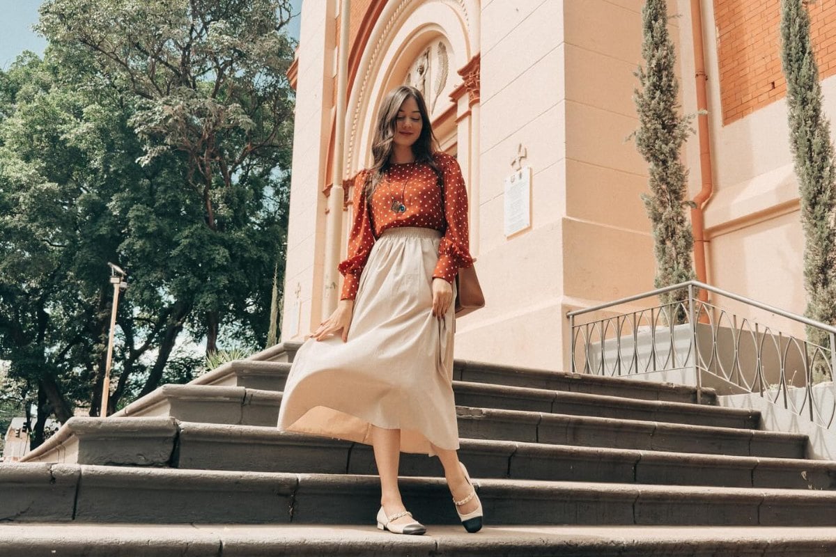 Uma mulher está descendo escadas ao lado de uma igreja. Ela veste uma blusa de manga longa vermelha com bolinhas brancas, e uma saia longa bege. Seus cabelos são longos e soltos. Ela sorri levemente, e carrega uma bolsa marrom. Árvores verdes estão ao fundo.