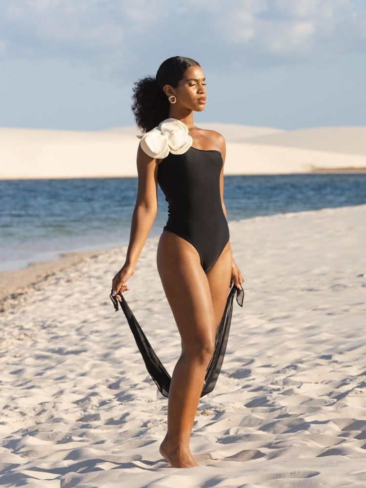 Mulher de pele negra na praia, usando maiô preto com máxi flor branca no ombro