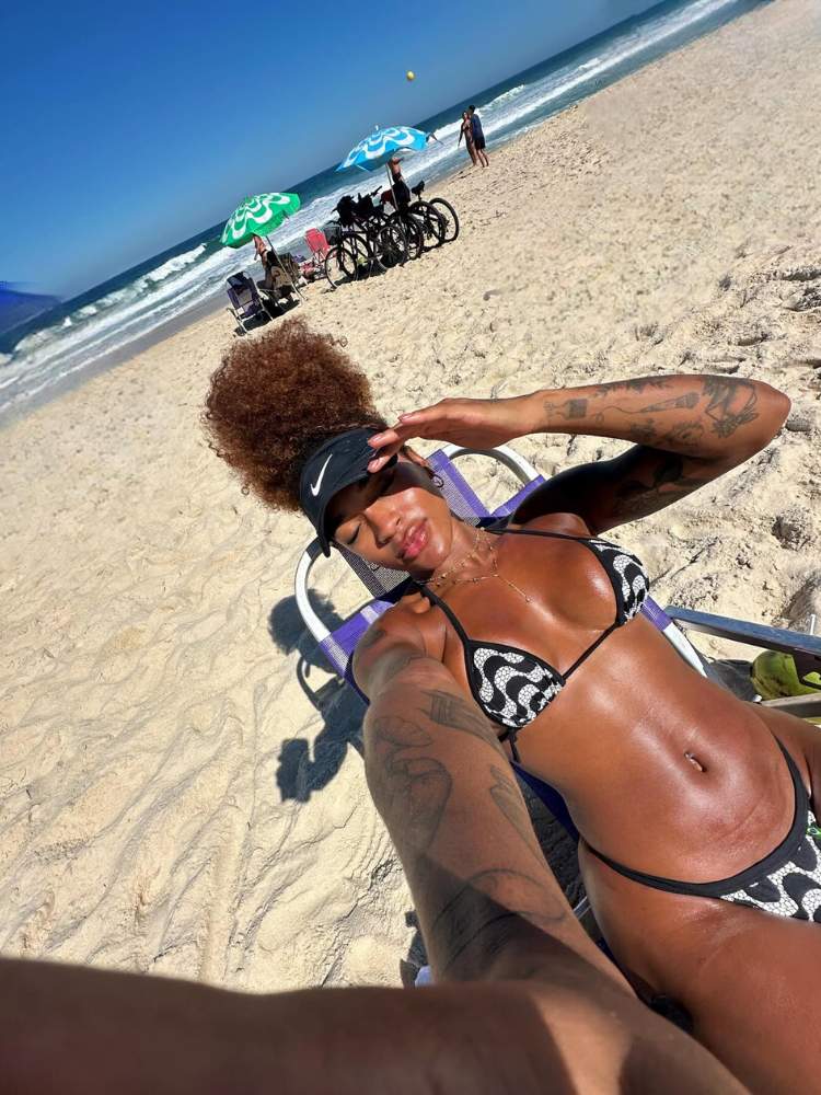 Patrícia Ramos tomando sol na areia da praia, usando viseira preta e biquíni branco e preto com estampa de calçada