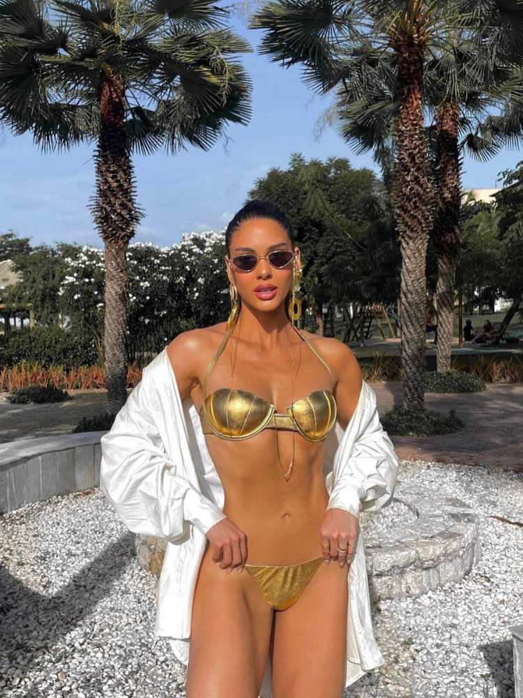 Mulher de pele clara usando biquíni dourado, com o top em formato de concha, além de saída de praia branca 