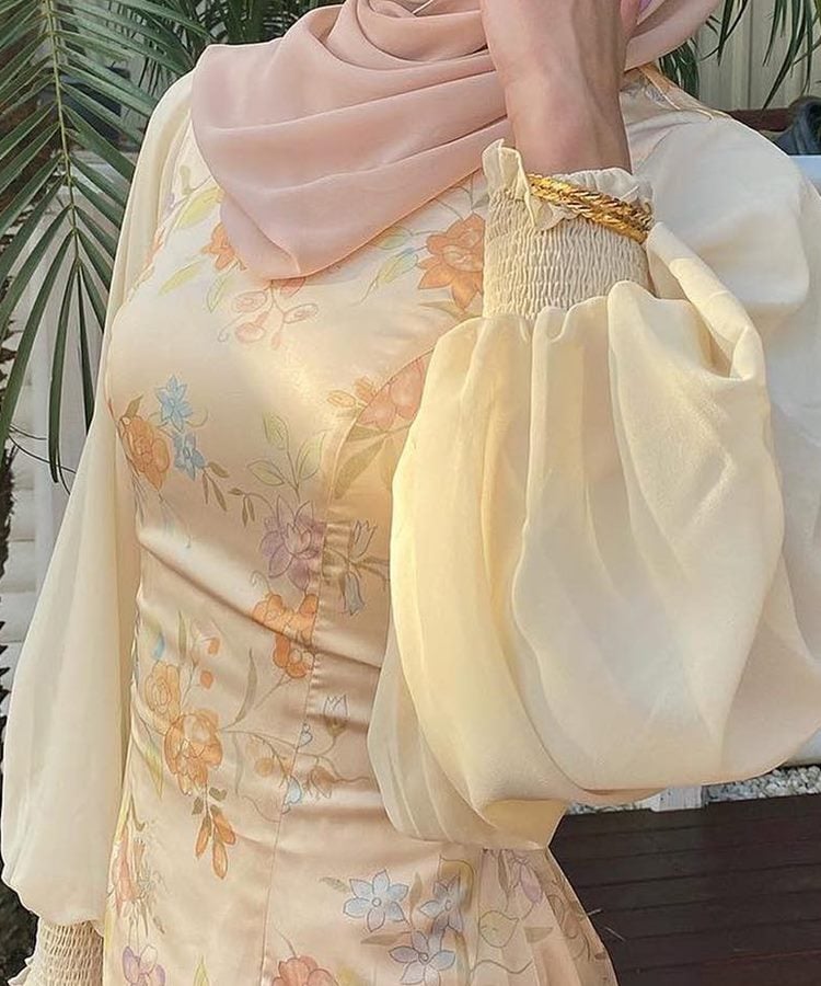 Uma mulher veste um vestido bege com estampa floral e mangas bufantes transparentes. Ela usa um hijab rosa claro e uma pulseira dourada. Este estilo delicado e sofisticado é um exemplo de moda modesta.