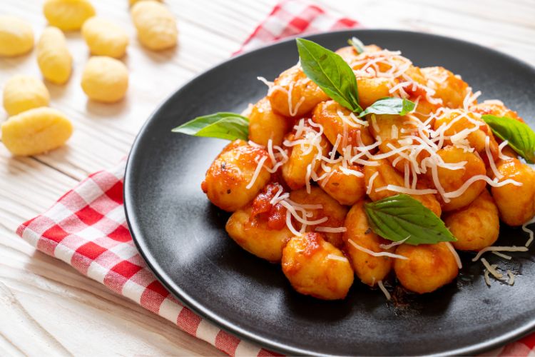 A imagem mostra um prato de nhoque coberto com molho de tomate e queijo ralado, decorado com folhas de manjericão fresco. O nhoque está servido em um prato preto sobre uma mesa de madeira, com um pano de prato xadrez vermelho e branco ao lado. Ao fundo, há algumas peças de nhoque não cozidas.