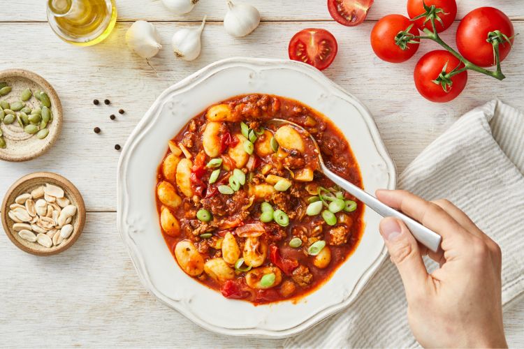 A imagem mostra um prato de nhoque coberto com um rico molho de tomate e pedaços de carne, decorado com cebolinha e sementes. Uma mão segura uma colher, pronta para saborear o prato. Ao redor, há ingredientes frescos como tomates, alho e azeite. Este prato é perfeito para a simpatia do Nhoque da Fortuna, trazendo sabor e tradição à mesa.