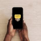 A foto mostra duas mãos segurando um smartphone. A tela do smartphone exibe uma interface de reprodução de música, com um fundo preto e um ícone de áudio amarelo. O texto visível na tela diz: "Pra Você" seguido por "Filipe Ret, M...". O fundo da imagem é uma superfície de madeira clara.