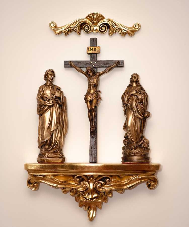 Nesta imagem, um oratório em casa é composto por uma prateleira dourada ornamentada. No centro, há um crucifixo com a inscrição "INRI". Ao lado, estão duas estátuas douradas, uma de cada lado, representando figuras religiosas. Acima do crucifixo, há um detalhe decorativo dourado na parede.