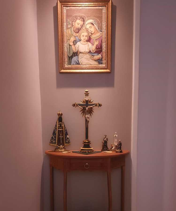 Esta imagem mostra um oratório em casa, localizado em um canto de parede. Há uma mesa de madeira com um crucifixo centralizado. À esquerda, uma imagem de Nossa Senhora Aparecida, e à direita, pequenas estátuas de santos. Acima, na parede, há um quadro com uma imagem da Sagrada Família emoldurada.