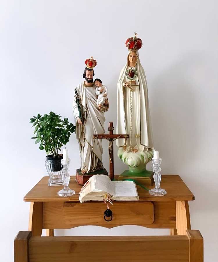 Este oratório em casa está sobre uma mesa de madeira. No centro, há um crucifixo pequeno. À esquerda, uma estátua de São José segurando o Menino Jesus, e à direita, uma estátua de Nossa Senhora de Fátima. Ambas as estátuas têm coroas vermelhas. Entre elas, há um livro aberto e dois castiçais de vidro com velas brancas. Um vaso com uma planta verde está à esquerda.