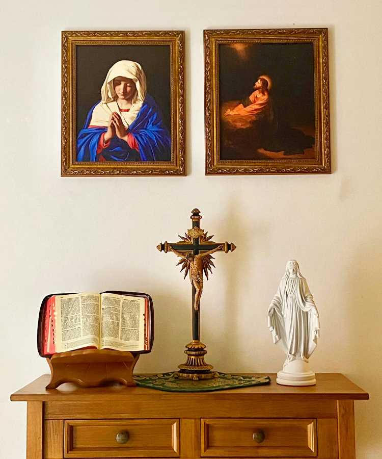 O oratório em casa está sobre uma cômoda de madeira. No centro, há um crucifixo ornamentado. À esquerda, um livro aberto em um suporte, e à direita, uma estátua branca de um santo. Na parede, dois quadros emoldurados com imagens religiosas.