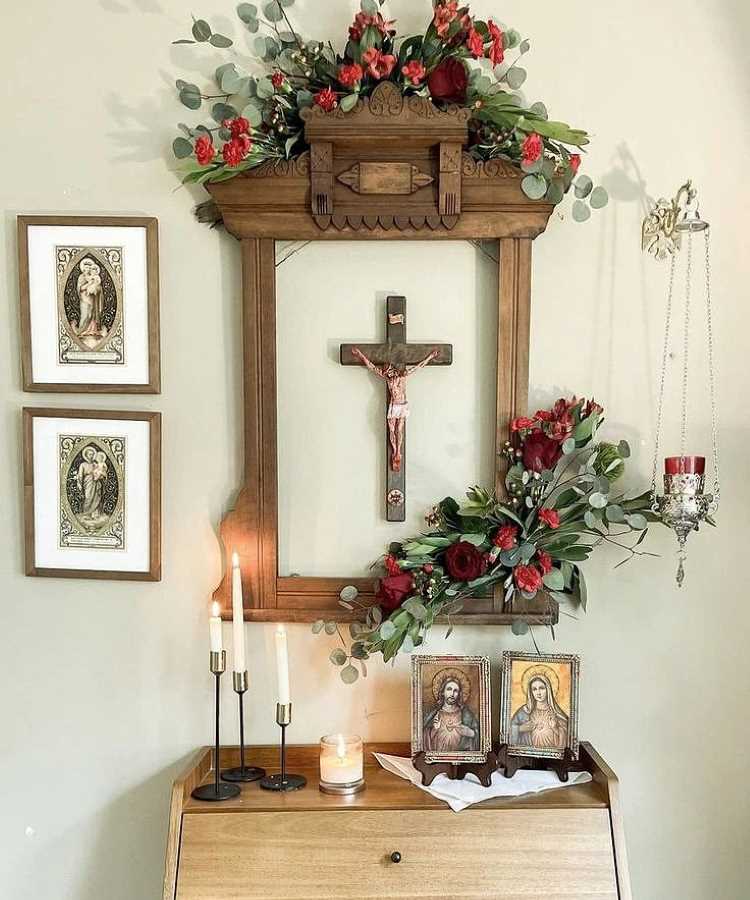 A imagem mostra um oratório em casa com um crucifixo central emoldurado por madeira decorativa. No topo, há arranjos de flores vermelhas e folhas verdes. Abaixo, duas imagens religiosas emolduradas estão sobre uma mesa de madeira clara. Ao lado, três velas em castiçais pretos e uma vela menor acesa. Na parede, dois quadros com imagens religiosas.