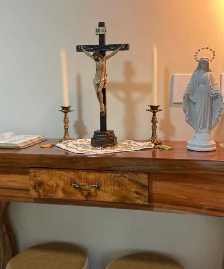  A imagem mostra um oratório em casa sobre uma mesa de madeira. No centro, há um crucifixo com a inscrição "INRI". Ao lado, duas velas em castiçais dourados e uma estátua branca de Nossa Senhora. À esquerda, um livro aberto sobre a mesa, que está coberta por uma toalha decorativa.