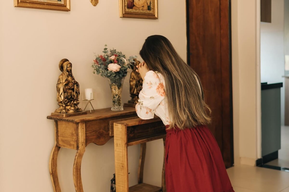 A imagem mostra uma mulher de cabelos longos e castanhos, vestindo uma blusa branca com estampas florais e uma saia vermelha. Ela está de pé, inclinada sobre uma mesa de madeira, com as mãos juntas em posição de oração. Na mesa, há uma estátua dourada de uma figura religiosa e um vaso de vidro com flores coloridas. A parede ao fundo é clara, decorada com quadros emoldurados. O ambiente parece ser uma sala de estar ou um espaço de oração.