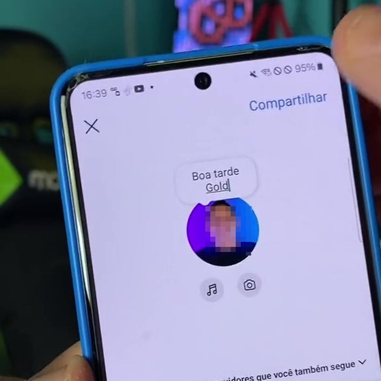 A imagem mostra a tela de um smartphone exibindo um perfil no Instagram. No centro da tela, há uma mensagem de saudação que diz "Boa tarde Gold". Abaixo da mensagem, há a foto de perfil do usuário, que está pixelada para preservar a privacidade. Ao redor da foto de perfil, há ícones de música e câmera. No canto superior direito, há a opção "Compartilhar". A mão de uma pessoa segura o telefone, que está em uma capa azul. Ao fundo, é possível ver um ambiente desfocado com uma cadeira e um objeto decorativo.